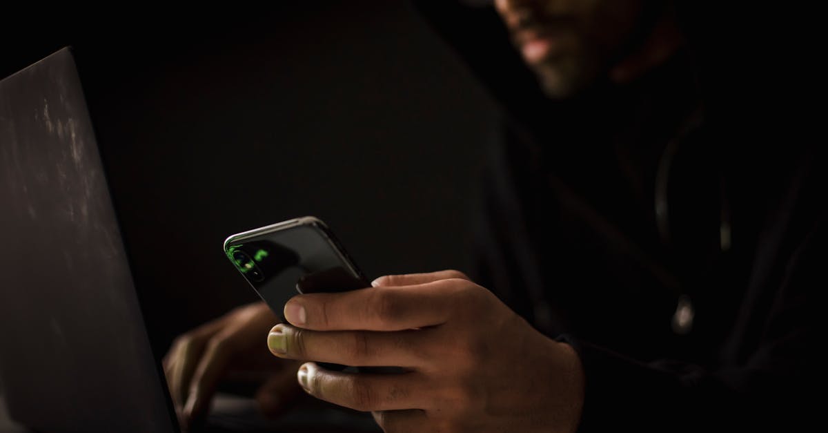 Crop anonymous ethnic male cyber spy with cellphone and netbook hacking system in evening